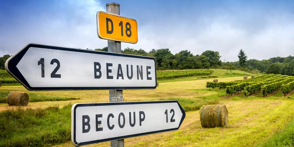 Una señal de carretera francesa que apunta a Beaune en una dirección y a Becoup en otra, con pacas de heno en el fondo
