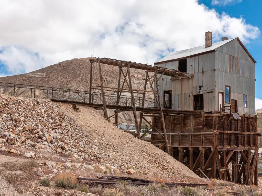 Parque Minero Histórico Tonopah.  Edificio de chapa corrugada de madera oscura y gris sobre el pozo de la mina bajo un paisaje azul de nubes.  Decapado beige o montón de roca estéril.