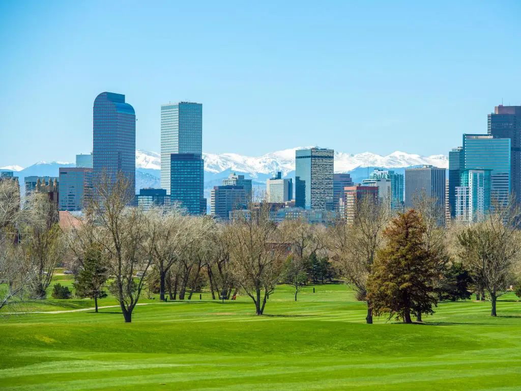 Denver, Colorado, Estados Unidos, tomada en un día soleado con el horizonte de la ciudad en la distancia y las Montañas Rocosas Nevadas, con árboles y hierba en primer plano.