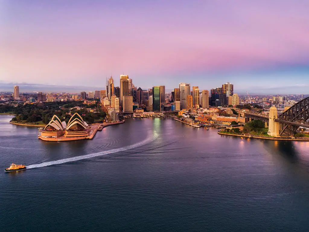 Puerto de Sydney, Australia, con un amanecer de color rosa sobre los puntos de referencia del CDB de la ciudad de Sydney en las costas del puerto de Sydney tocados por el arco de acero del puente del puerto con el ferry de pasajeros en ruta al muelle.