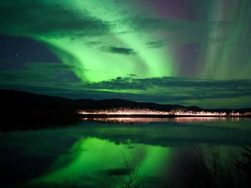 Estrellas del cielo nocturno, nubes y auroras boreales sobre la carretera rural en la orilla del lago