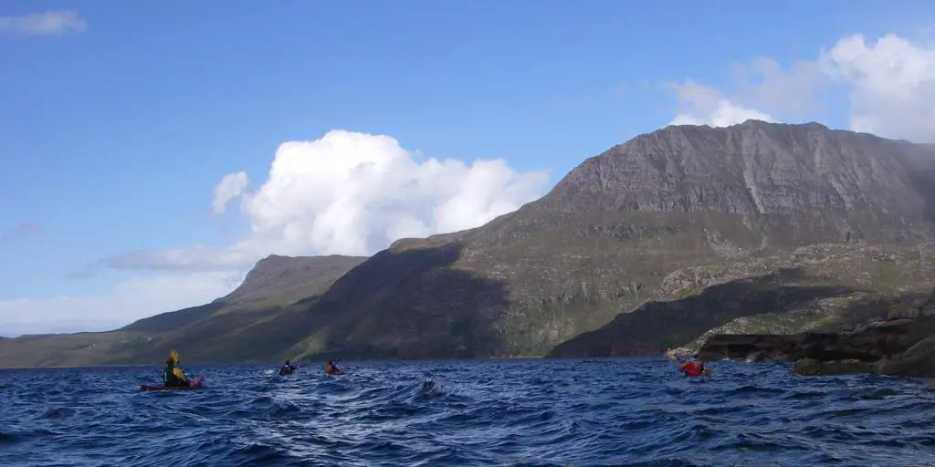 La punta de un kayak en el agua, con otros kayaks y montañas al fondo