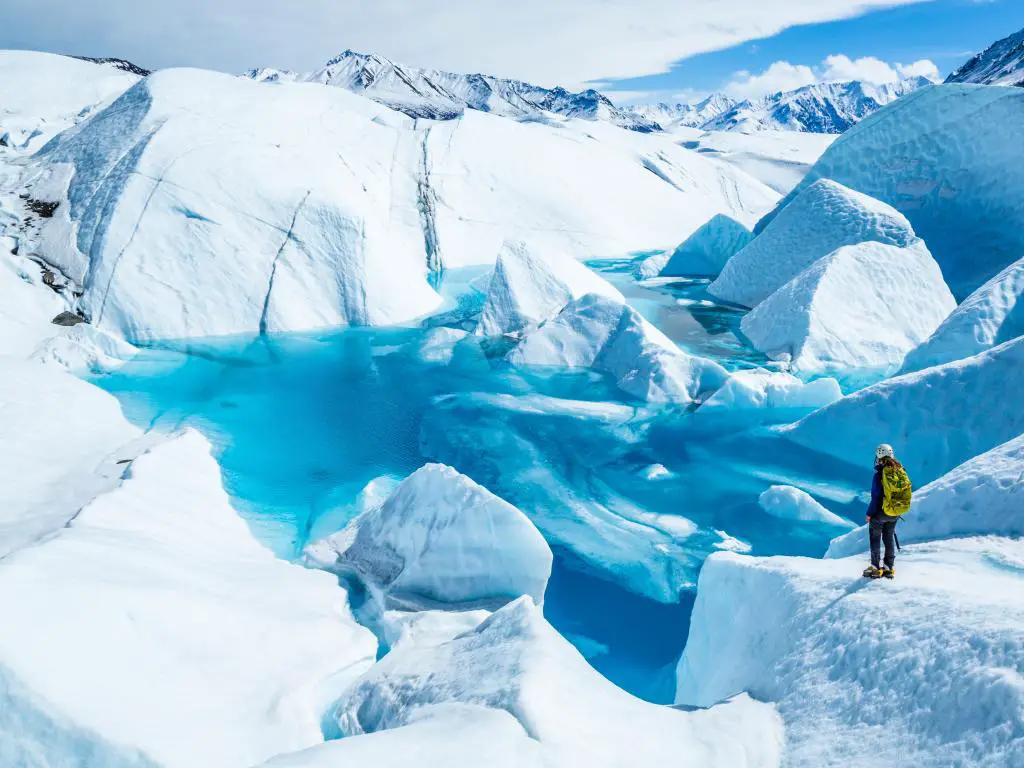De pie cerca del borde de una gran piscina azul en la parte superior del glaciar Matanuska.  Una mujer joven que sostiene un piolet con una mochila y un casco mira hacia el lago.
