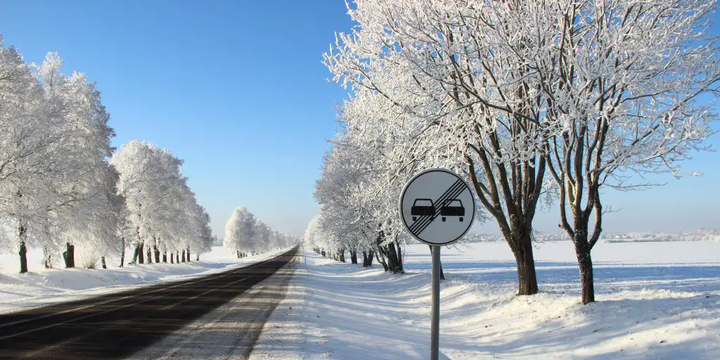 Señal de tráfico prohibido adelantar al lado de una carretera principal vacía, con cielo azul y nieve en el suelo y en los árboles