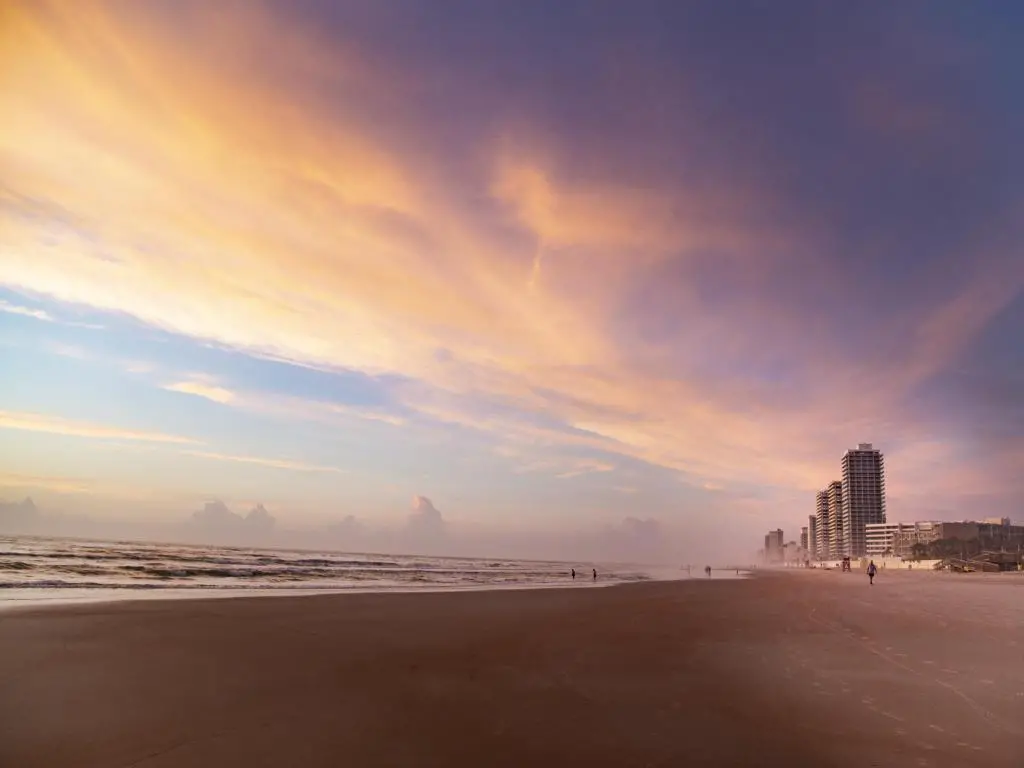 Daytona Beach, Florida, EE. UU., con una vista impresionante de una puesta de sol rosa y una vista de la playa con edificios a lo lejos.