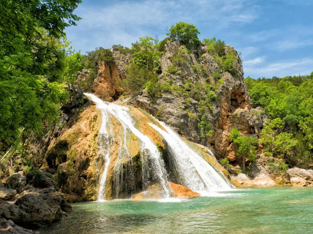 Turner Falls, Oklahoma, EE.UU. con agua en cascada sobre rocas en una piscina natural en un día soleado rodeado de árboles.