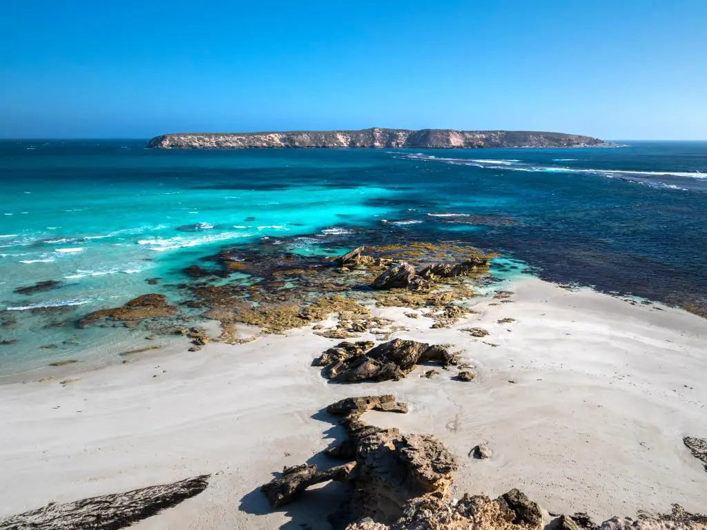 Parque Nacional Coffin Bay, Península de Eyre, Australia Meridional con rocas y arena blanca en primer plano, un mar azul y una isla a lo lejos en un día despejado.