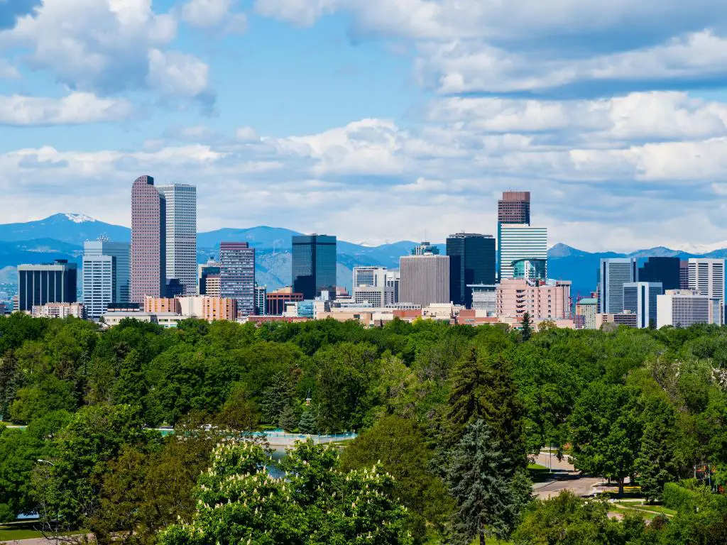Denver, Colorado, Estados Unidos, con los rascacielos de la ciudad en la distancia y las montañas más allá, además de árboles en primer plano en un día soleado.