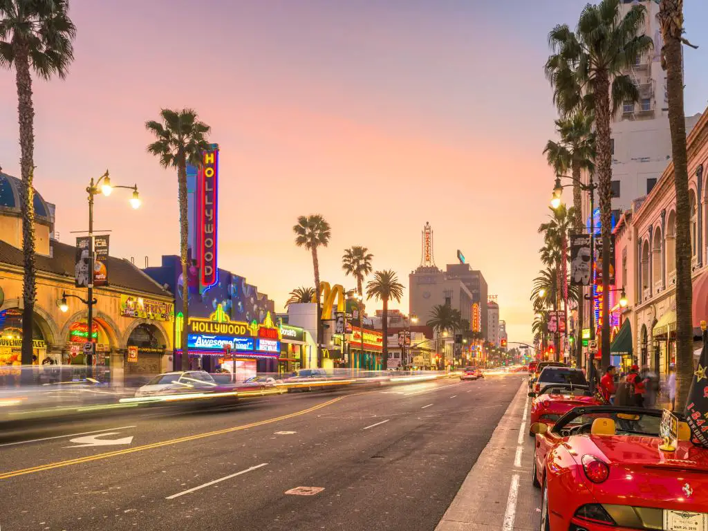 Los Ángeles, California, EE.UU. con tráfico y autos deportivos en Hollywood Boulevard al atardecer con palmeras y edificios icónicos en la distancia.