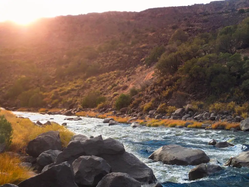 Río Grande, Nuevo México, EE. UU. Tomado como una vista del río a orillas del río cuando el sol se hunde está a punto de sumergirse debajo del borde del desfiladero mientras el río balbucea a través de las rocas en el frente.