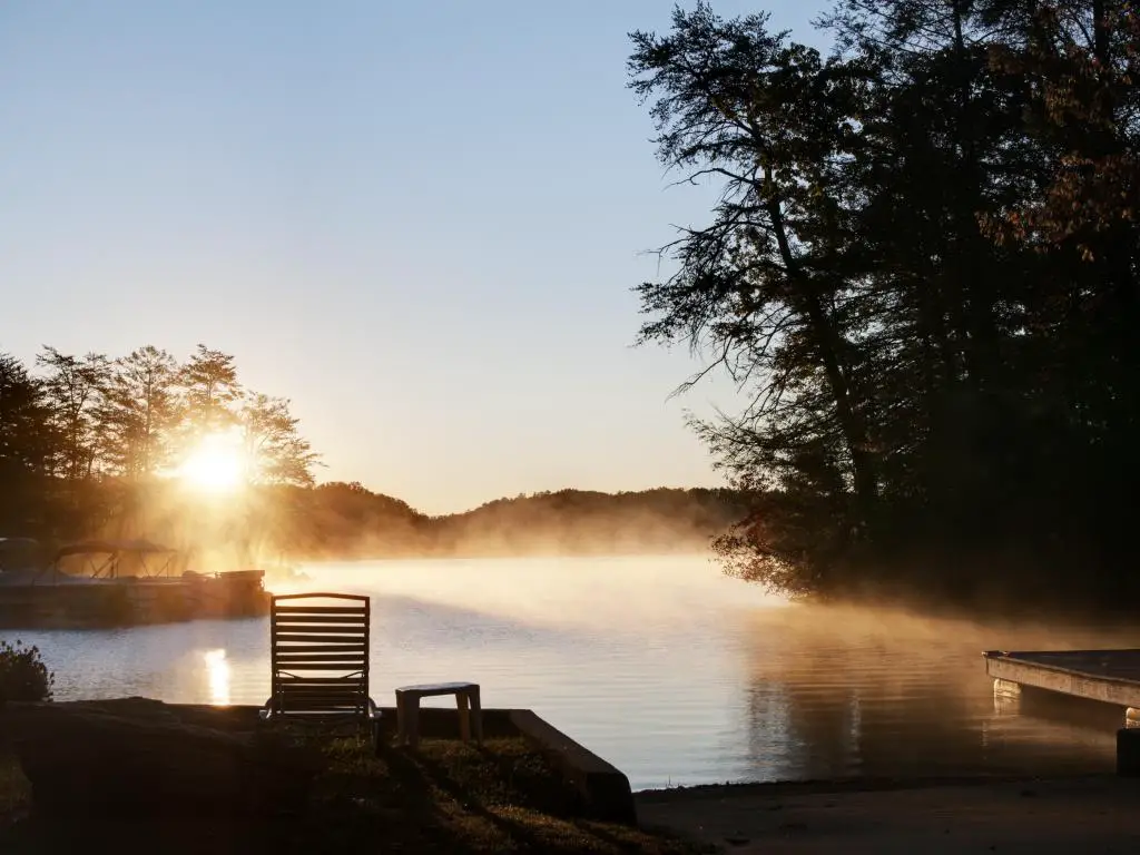 Lake Lure, Carolina del Norte, EE.UU. con el sol saliendo sobre el lago y la niebla a lo lejos, árboles altos en las sombras.