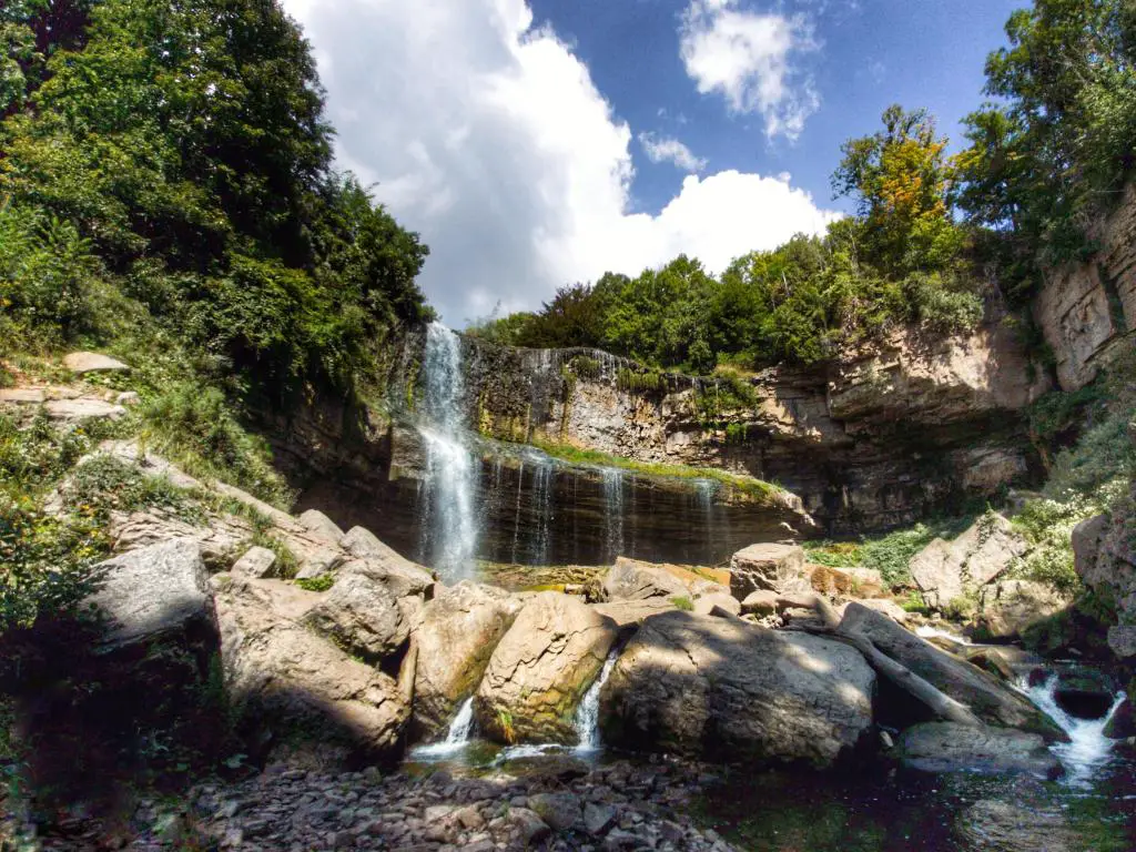 Webster Falls, Hamilton, Ontario, Canadá, tomada en un día soleado con la cascada rodeada de follaje verde y aterrizando en las rocas de abajo.