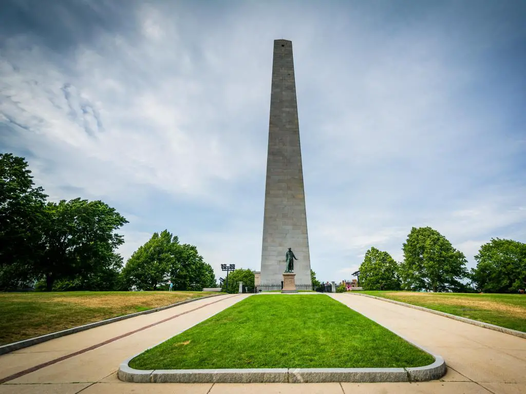 El Bunker Hill Monument, Charlestown, Boston, Massachusetts, EE.UU., tomado en un día soleado con hierba en primer plano y árboles en la distancia.