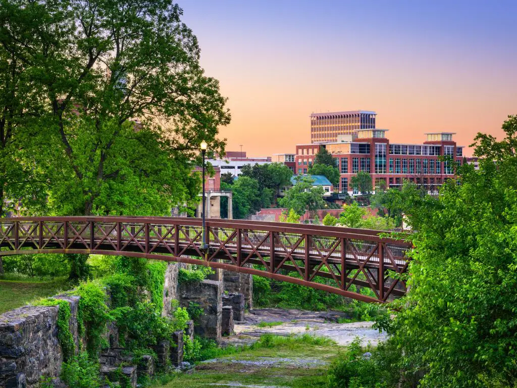El horizonte y el parque del centro de Columbus, Georgia, Estados Unidos, con un puente que cruza un río y muchos árboles y vegetación en primer plano al atardecer.