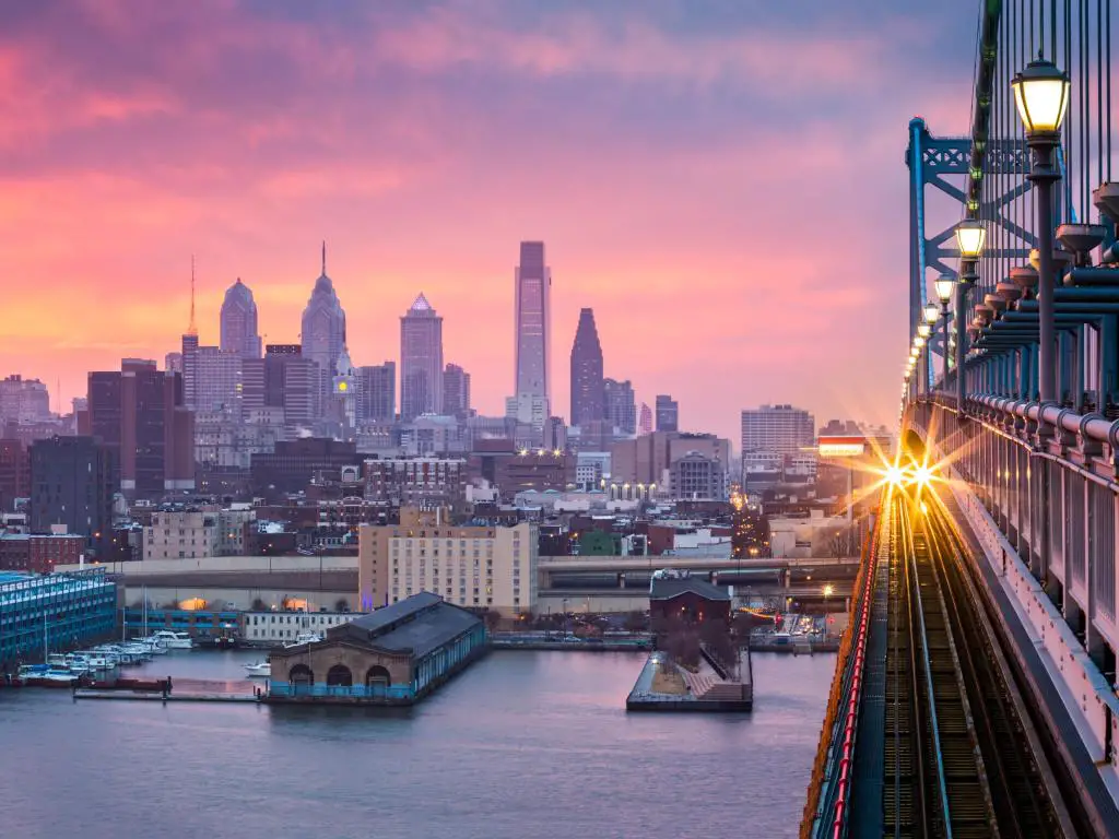 Filadelfia, EE.UU. con una vista panorámica del horizonte de la ciudad en la distancia bajo una puesta de sol púrpura nebulosa con un tren entrante cruzando el puente Ben Franklin.