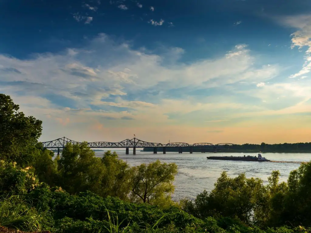 Vicksburg, Mississippi, EE.UU. con un barco en el río Mississippi, cerca del puente de Vicksburg al atardecer, árboles en primer plano.