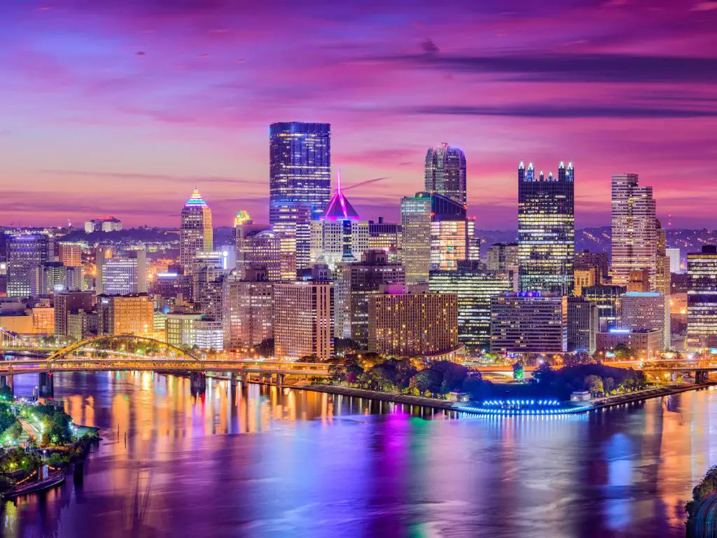 Pittsburgh, Pensilvania, EE.UU. con el horizonte de la ciudad en la distancia tomada al atardecer con un cielo púrpura y la ciudad reflejada en el agua en primer plano.