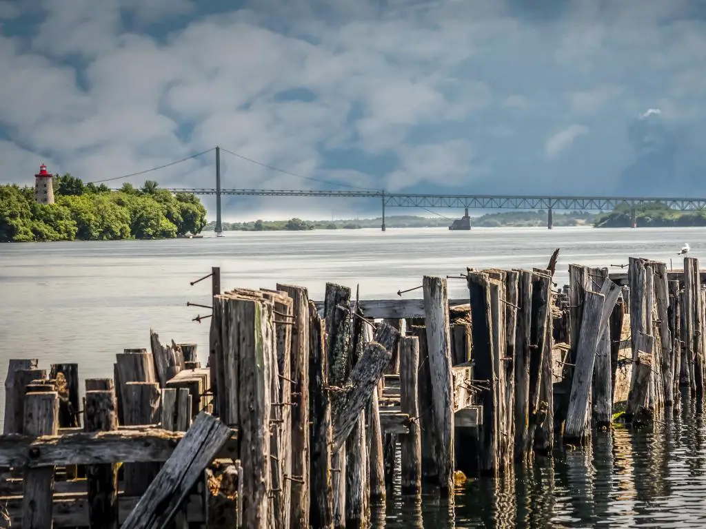 Puente internacional Seaway, Canadá/EE.UU. con pilotes antiguos de un antiguo puerto con un faro de 1800 y el puente que cruza el río San Lorenzo en un día nublado.