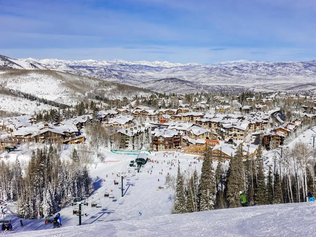 Deer Valley Ski Resort, Utah con un telesilla, pistas de esquí en primer plano y la estación de esquí antes de las montañas cubiertas de nieve en la distancia en un día soleado.