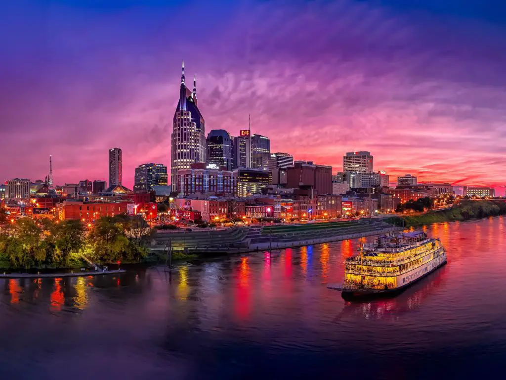 Cruceros fluviales iluminados pasando por edificios de gran altura en la orilla del río, con luz de puesta de sol de color rosa oscuro y azul