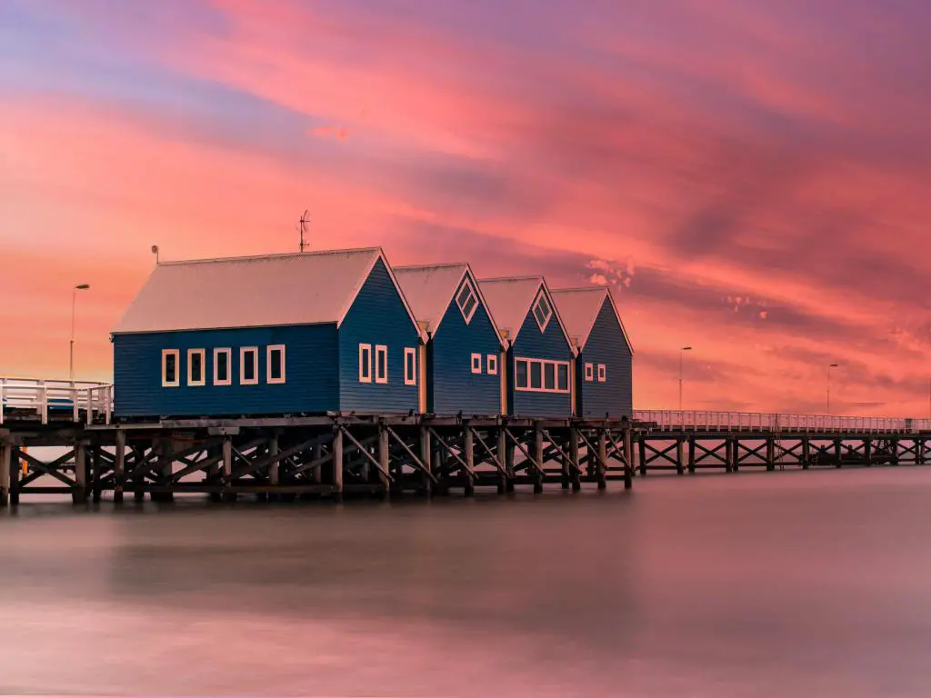 El embarcadero de madera se extiende en la distancia sobre el océano con tres edificios de madera en primer plano, bajo un cielo rosa vivo al atardecer.