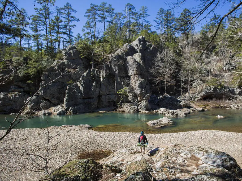 Montañas Ouachita, Arkansas con un excursionista caminando hacia un río con formaciones rocosas en el fondo y árboles en la distancia. 