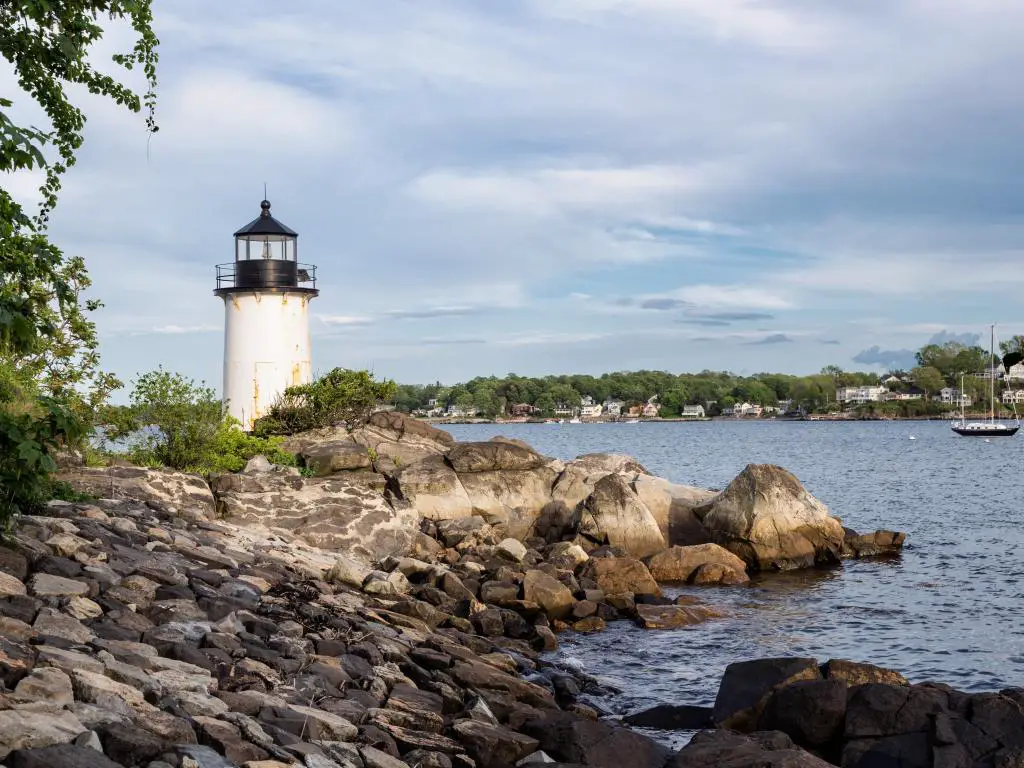 Salem, Massachusetts, EE.UU. con Winter Island Lighthouse detrás de rocas en primer plano, el mar que rodea y árboles y edificios en la distancia.
