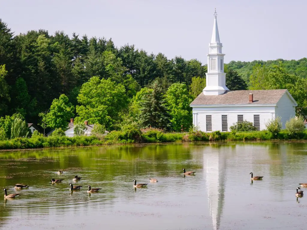Pequeña iglesia blanca junto a un lago con patos nadando, rodeada de árboles verdes