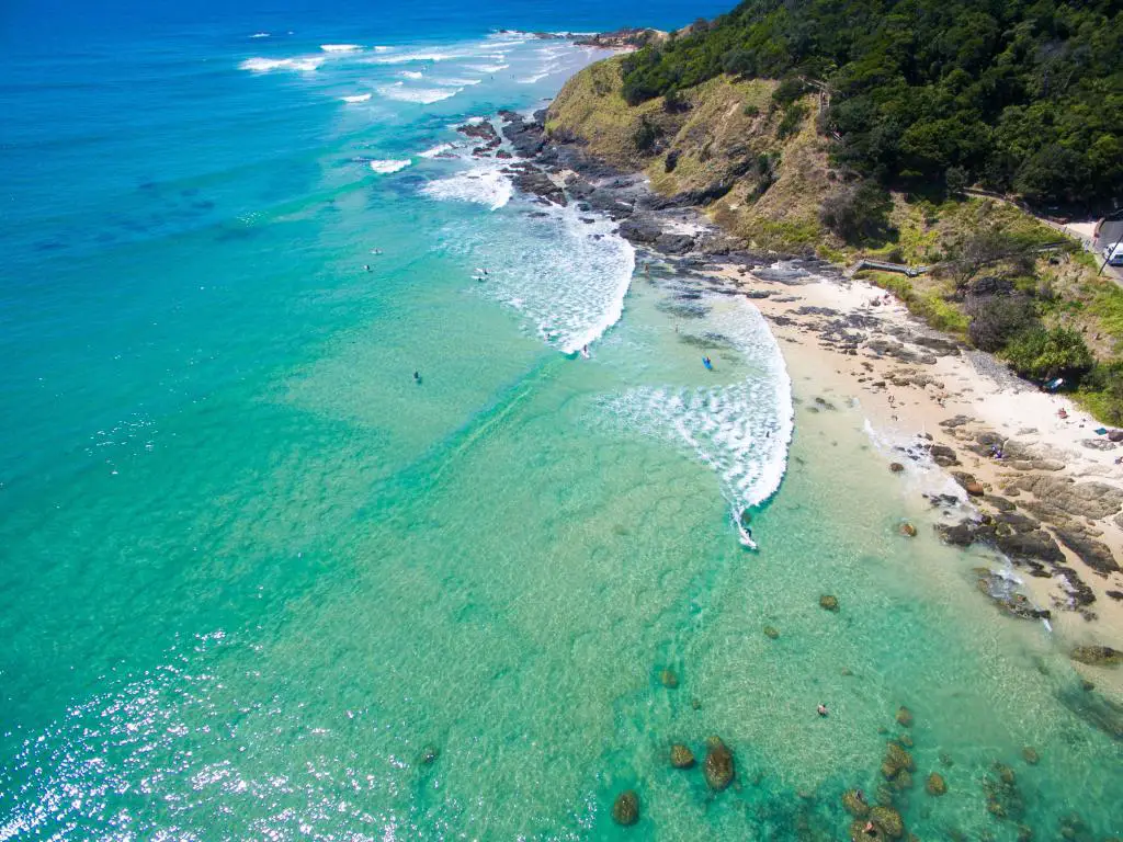 Vista aérea del mar azul claro con surfistas, con una playa de arena y rocas en la bahía y un acantilado que se eleva a un lado