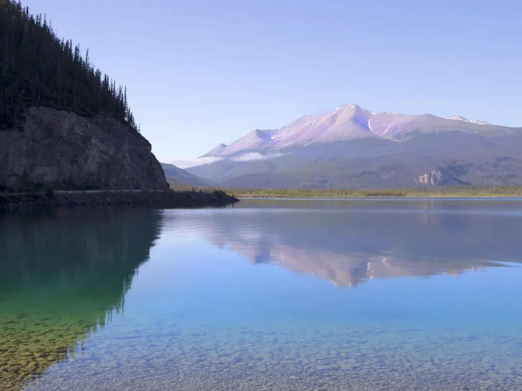Lago Muncho en Columbia Británica, Canadá, con aguas tranquilas en primer plano y montaña en la distancia tomada en un día soleado.
