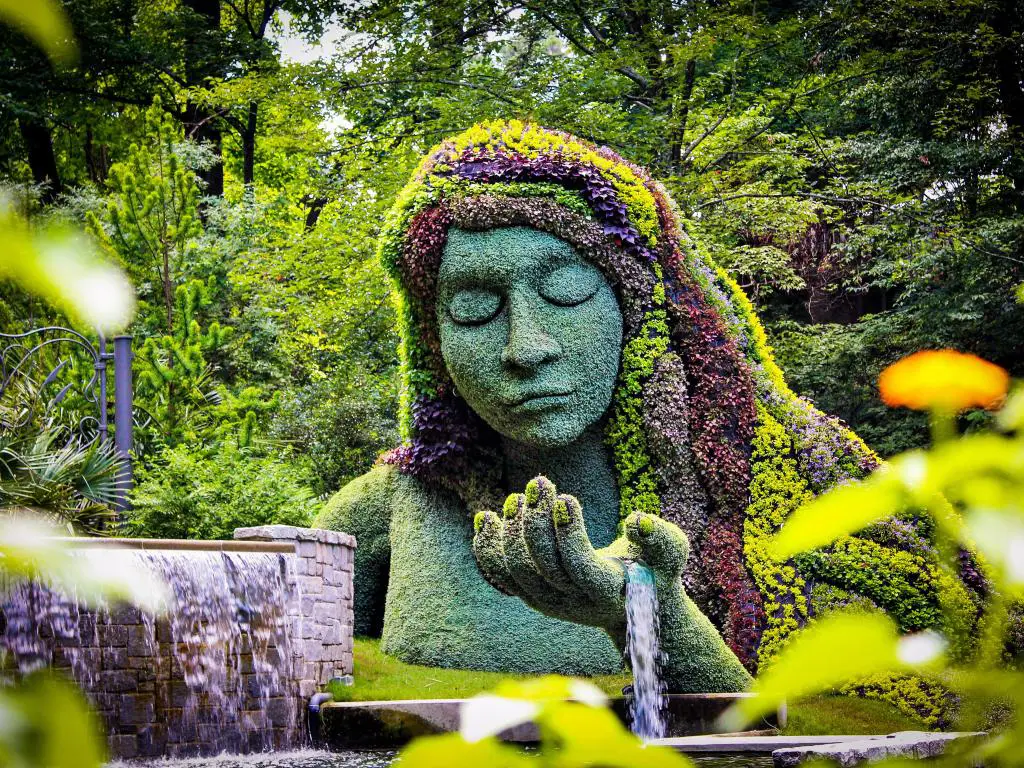 Escultura vegetal de la diosa de la tierra en los Jardines Botánicos de Atlanta.  El busto está cubierto de hierba mientras que su cabello está hecho de flores.  El agua fluye de su palma.  Sus ojos están cerrados.