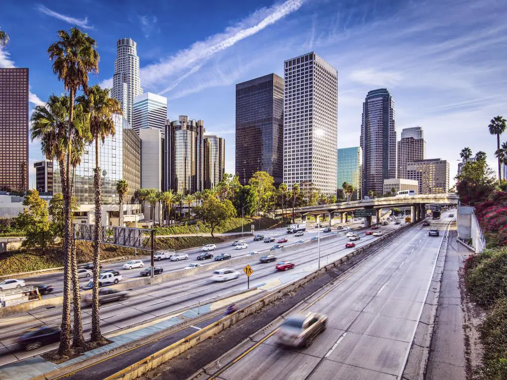 Los Ángeles, Estados Unidos, con el horizonte de la ciudad al fondo, tomado en un día claro y soleado, con tráfico que conduce a la ciudad y un par de palmeras en primer plano. 