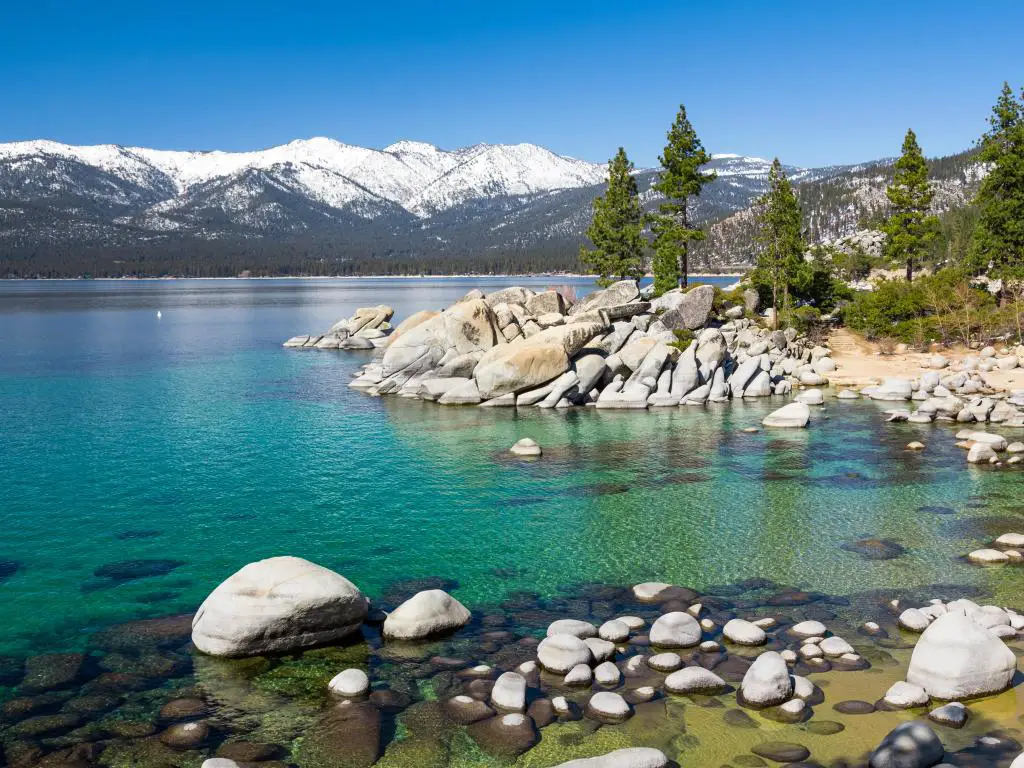 Lake Tahoe, Nevada y California, EE. UU., tomadas en un día claro y soleado con agua turquesa y piedras grandes en primer plano, algunos árboles que salpican la costa y montañas nevadas en la distancia. 
