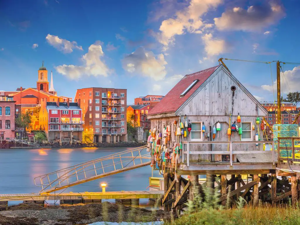 Embarcadero de madera y muelle adornado con boyas de colores, al lado del río con edificios bajos en la otra orilla