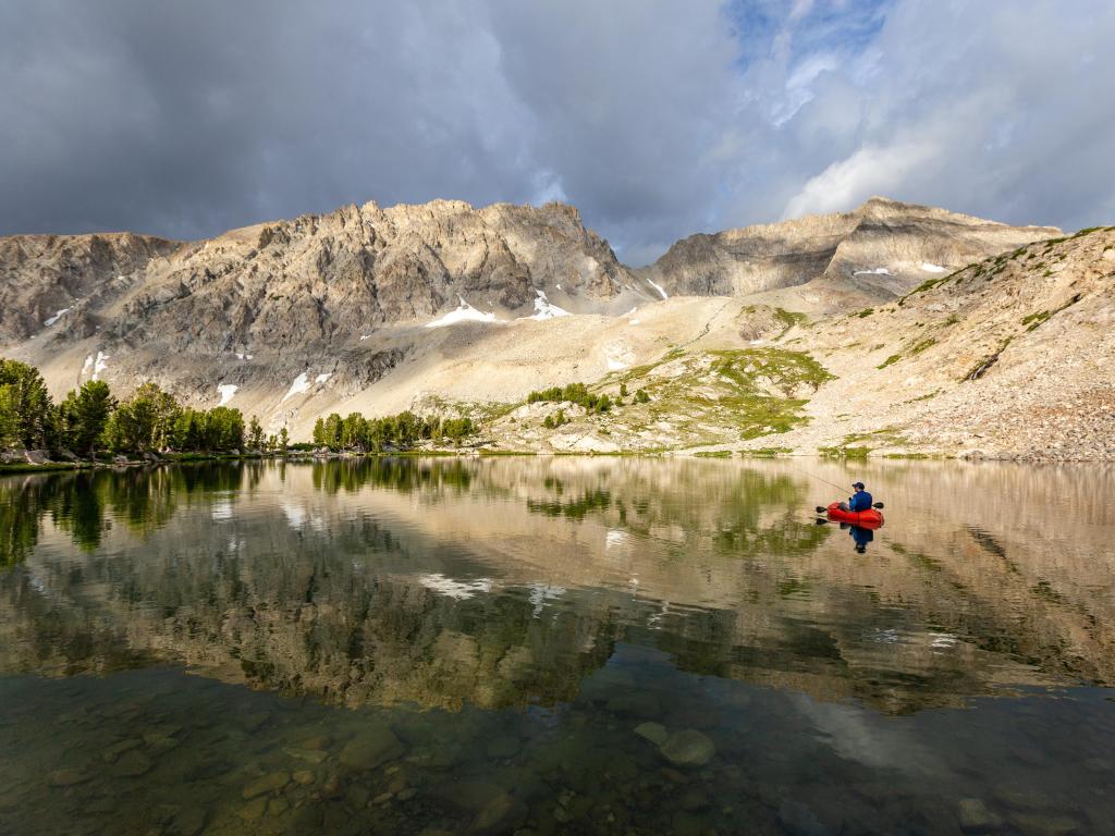 Sun Valley, Idaho, EE. UU. Con alguien pescando con mosca desde una balsa de carga en un lago alpino en primer plano, montañas y nubes arriba.