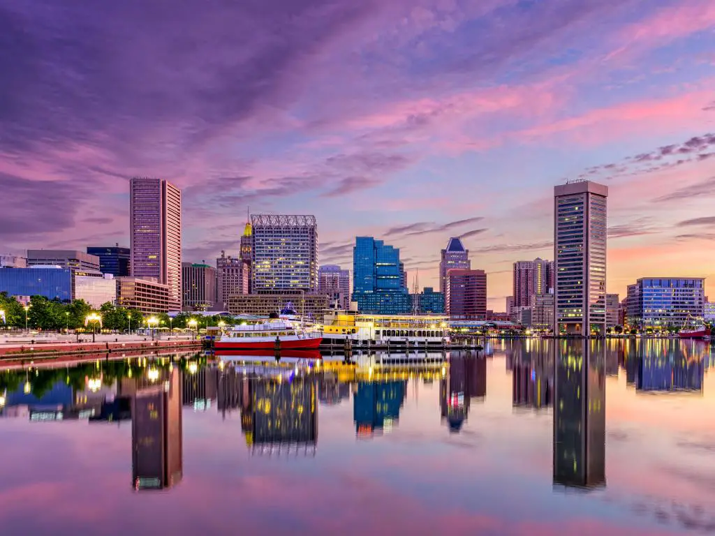 Baltimore, Maryland, EE.UU. con el horizonte de la ciudad en la distancia reflejándose en el agua en el puerto interior en primer plano y tomado después del atardecer con un cielo púrpura.
