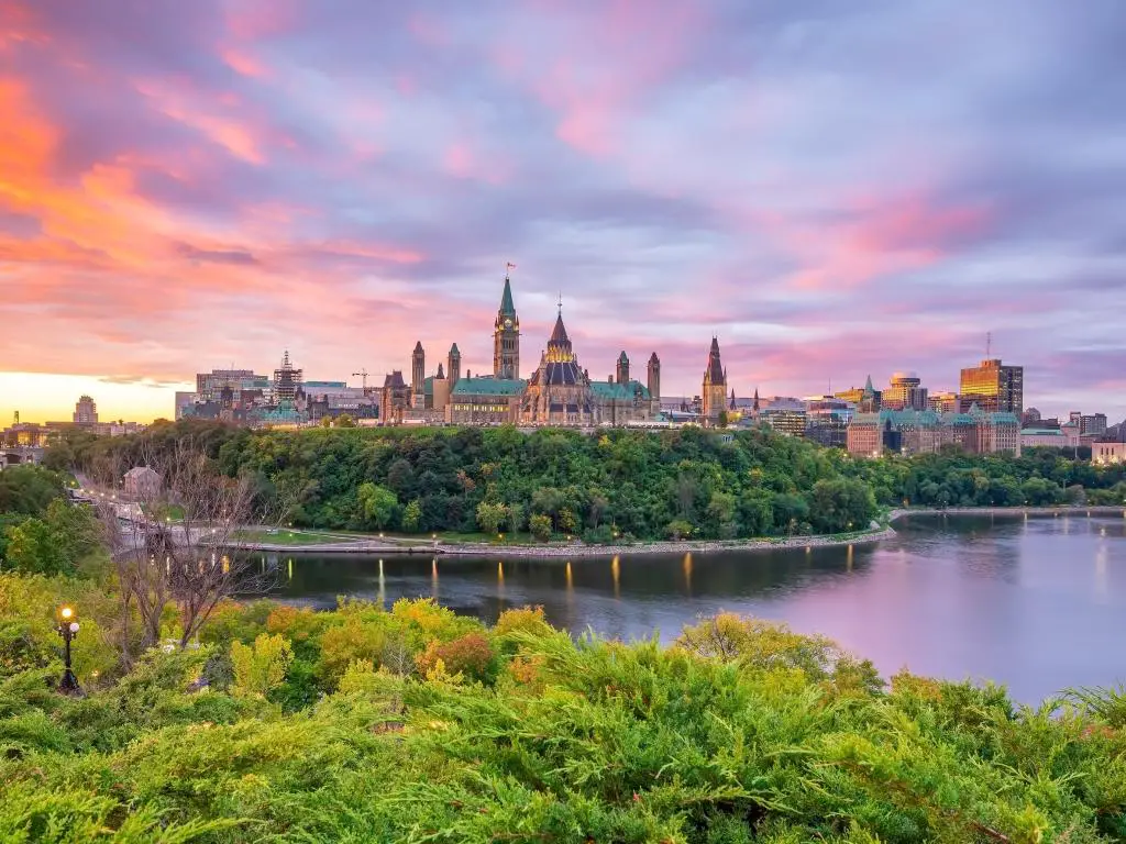 Parliament Hill en Ottawa, Ontario, Canadá, tomada al atardecer con árboles en primer plano y aguas tranquilas. 