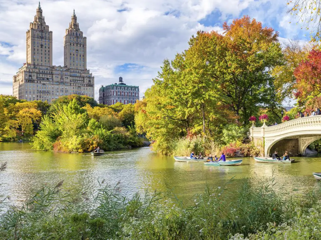 Central Park, Nueva York, Estados Unidos, tomada en otoño con hermosos colores de follaje, el puente en el borde y gente en botes en el lago y un edificio icónico en la distancia. 