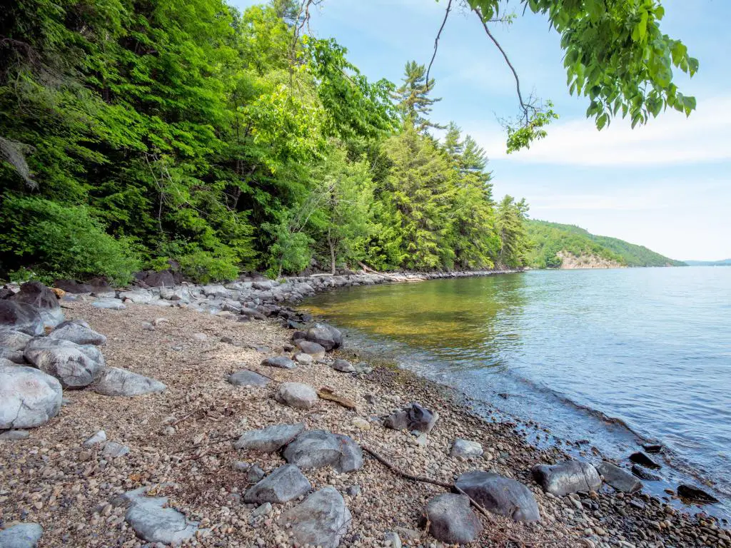 Lago Champlain, frontera entre EE.UU. y Canadá tomada en una bahía con una costa rocosa y árboles altos en la distancia en un día soleado.