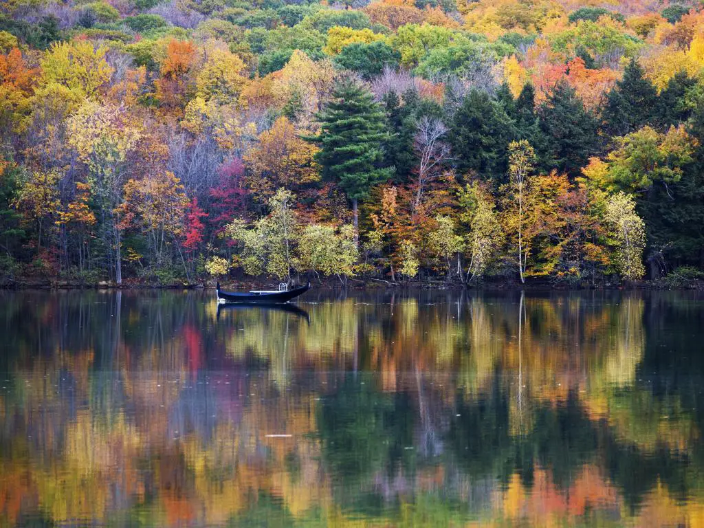 Parque Nacional Mont-Saint-Bruno, Canadá con árboles de otoño en varios colores que se reflejan en el lago en primer plano y un solo bote en el agua. 