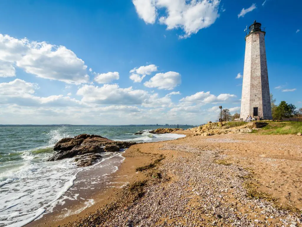 El faro de New Haven, en Lighthouse Point Park en New Haven, Connecticut, con arena en primer plano, mar suave y en un día claro y soleado.