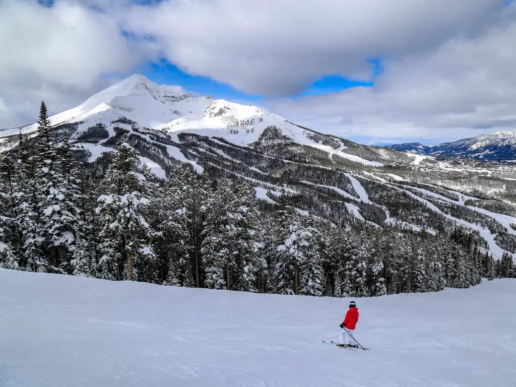 Big Sky, Montana, EE.UU. con un hombre en esquí alpino rojo y la montaña cubierta de nieve en la distancia tomada en un día soleado pero nublado.