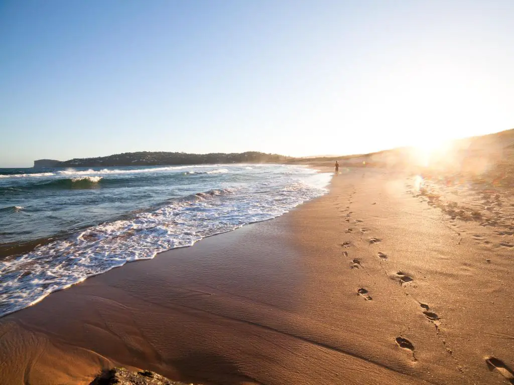 Palm Beach en Nueva Gales del Sur, Australia al atardecer con una figura caminando junto al mar y en la distancia dejando huellas en la arena. 