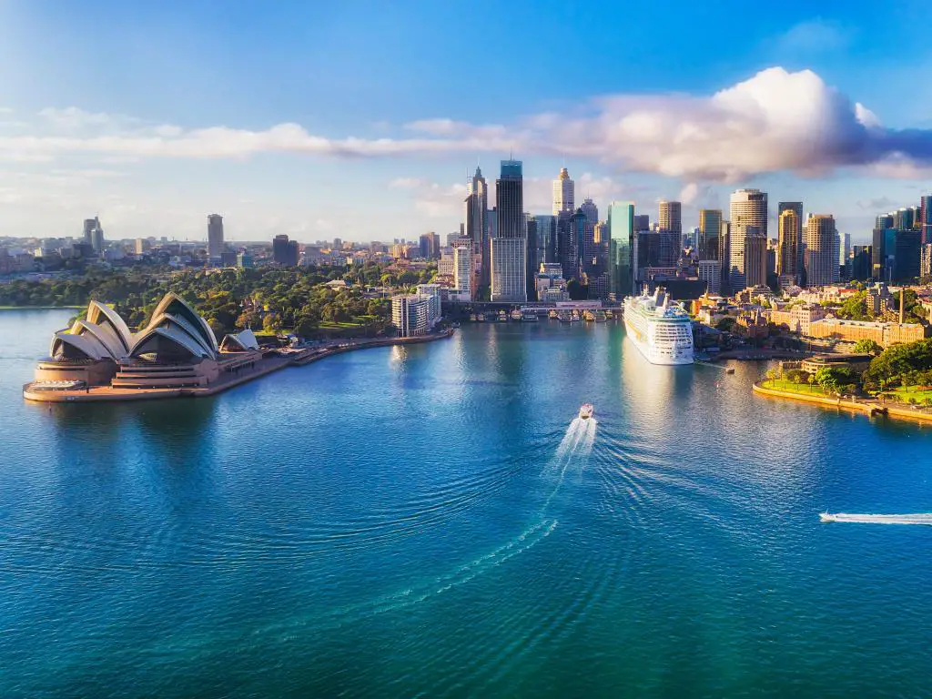 Puerto de Sydney, Australia, con los famosos monumentos de la ciudad al fondo, barcos en el mar en primer plano bajo el sol. 