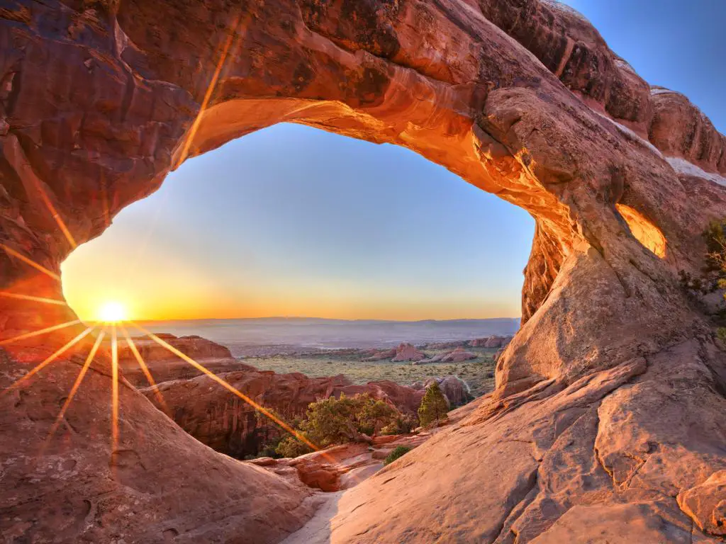 Vista a través del arco de roca roja natural al amanecer, a través de un amplio paisaje con rocas y árboles