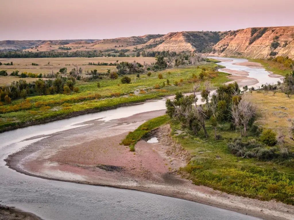 Parque Nacional Theodore Roosevelt, Dakota del Norte, con un río que atraviesa y una tierra verde plana a ambos lados que conduce a las montañas en la distancia al atardecer.