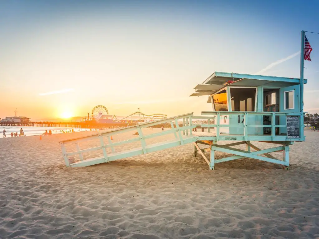Playa de arena con puesta de sol en el horizonte, con una cabina de salvavidas azul vibrante en primer plano y un parque de diversiones en el fondo