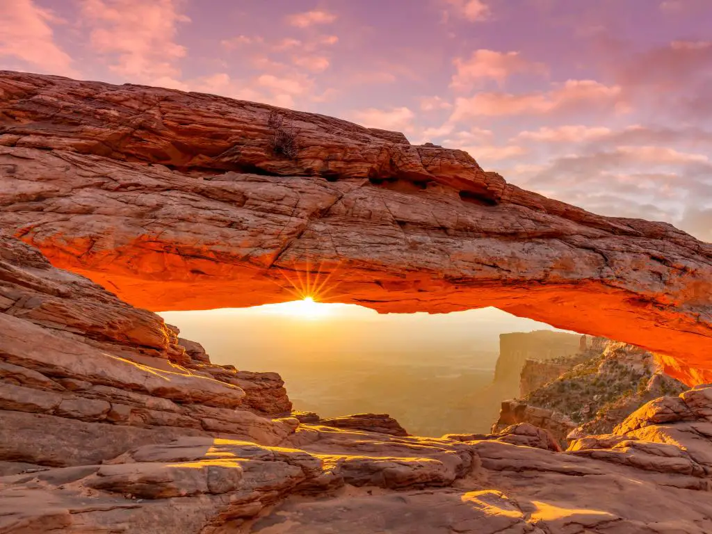 Salida del sol que envía un rayo de luz vibrante a través de una formación rocosa de color naranja en forma de arco