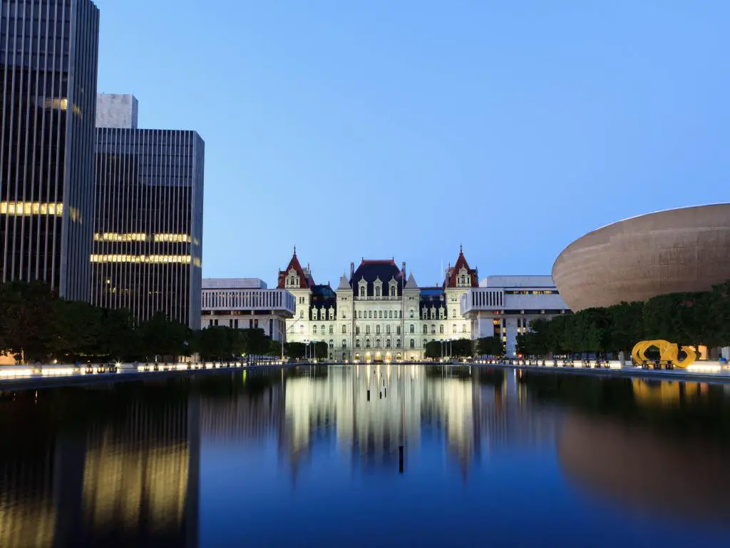 Histórico edificio blanco del Capitolio del Estado visto a través del agua del lago con edificios de gran altura a un lado y un edificio moderno al otro