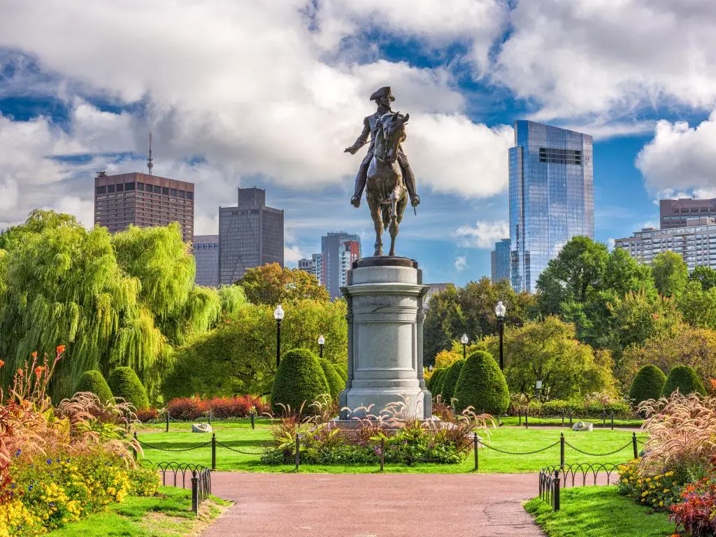 Boston, Massachusetts con el Monumento a George Washington en primer plano rodeado por el parque verde, con flores y el horizonte de la ciudad en la distancia en un día nublado pero soleado.
